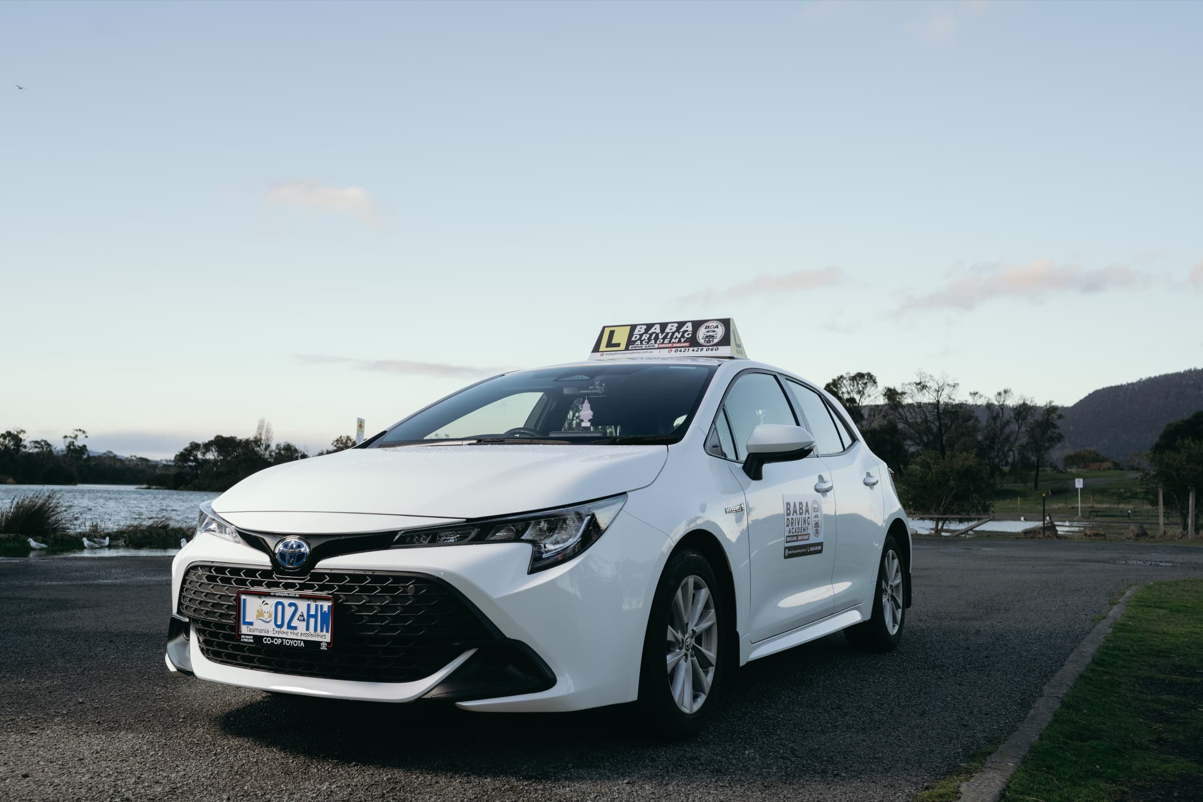 Baba Driving Academy's modern Toyota training vehicle parked by a lake in Hobart, Tasmania.